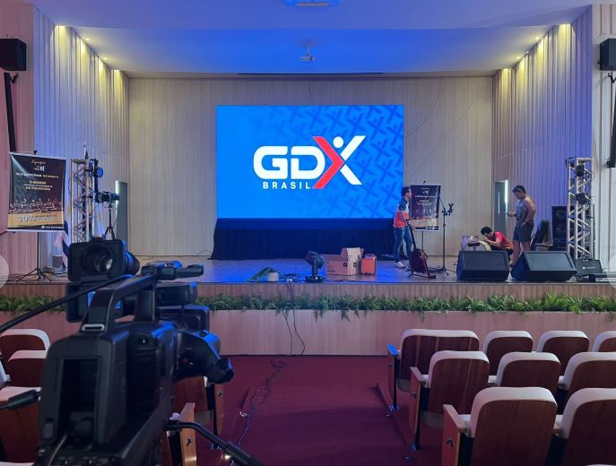 Auditório preparado pela GDX Brasil com grande painel de LED exibindo o logo GDX no palco, câmera de transmissão em primeiro plano e caixas de som instaladas.