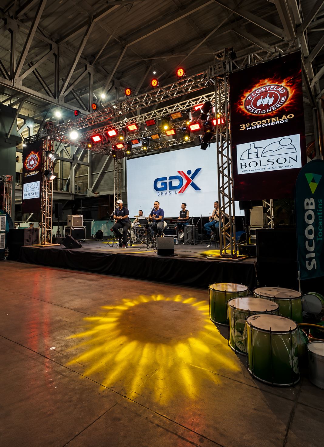 Palco do 3º Costelão Missioneiros montado pela GDX Brasil em pavilhão coberto, com treliças, iluminação cênica vermelha e amarela, painéis de LED laterais e telão central exibindo o logo GDX, com banda tocando ao vivo.
