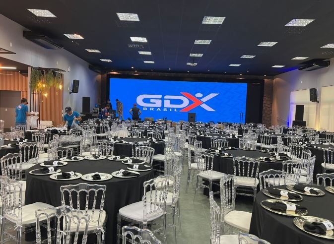 Salão de festas com mesas de gala postas e grande painel de LED exibindo o logo GDX Brasil ao fundo, com iluminação cênica e caixas de som instaladas.
